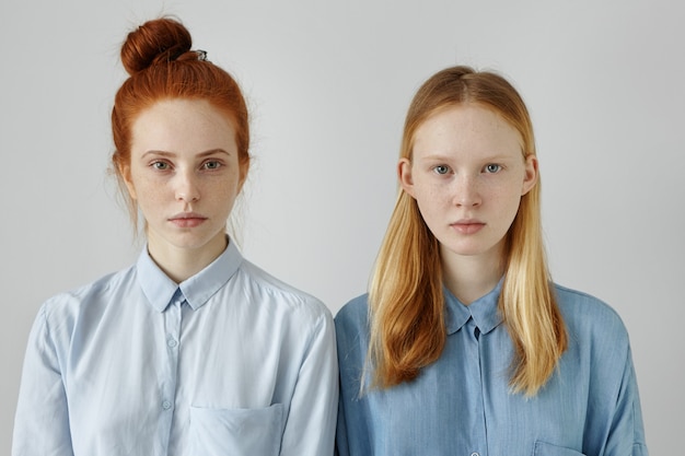 Gratis foto twee mooie vrouwen in shirts poseren op grijze muur. roodharige jonge vrouw met haarbroodje dat zich dicht bij haar blonde kleine zusje bevindt, beiden op zoek met zelfverzekerde uitdrukking