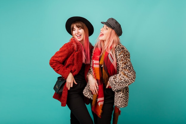 Twee mooie vrouwen in stijlvolle faux fur jassen en wollen sjaal poseren op turquoise muur