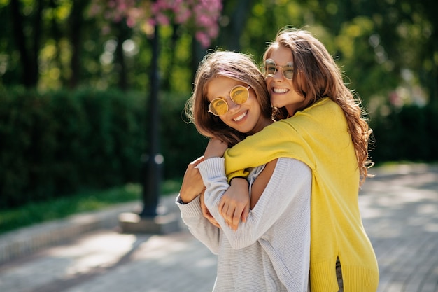 Twee ongelooflijke meisjes in heldere zonnebrillen en truien die elkaar op straat knuffelen, positieve stemming, echte emoties. Openluchtportret van twee jonge dames op straat