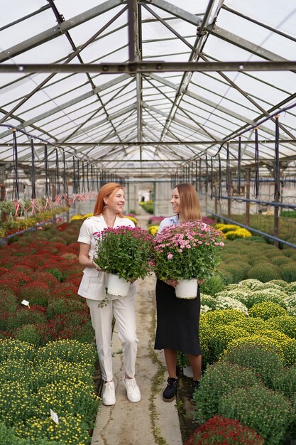 Twee schattige dames poseren met een bos roze chrysanten in een prachtig bloeiend groen huis met glazen dak.