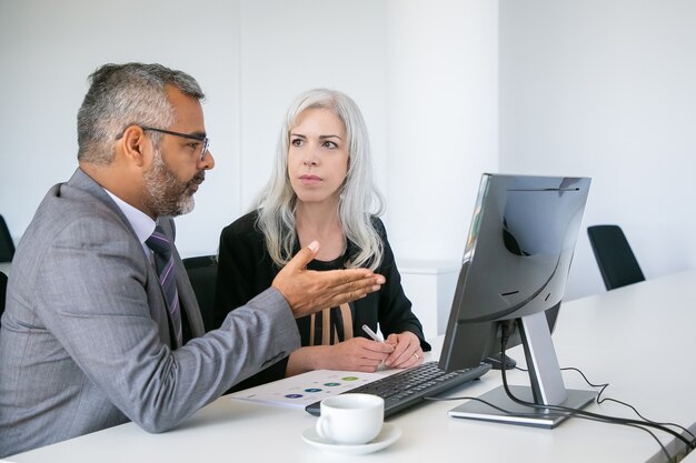 Twee serieuze managers kijken naar presentatie op pc-monitor, project bespreken, zittend aan een bureau met papieren diagram. Zakelijke communicatie concept