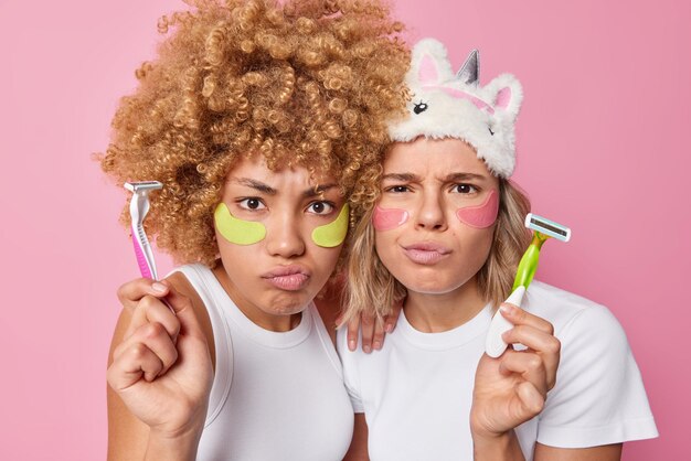 Twee serieuze vrouwen fronsen het gezicht met sombere uitdrukkingen passen hydrogelpleisters toe onder de ogen om de gevoelige huid te hydrateren, houden scheermesjes om te scheren gekleed in casual t-shirts geïsoleerd over roze muur