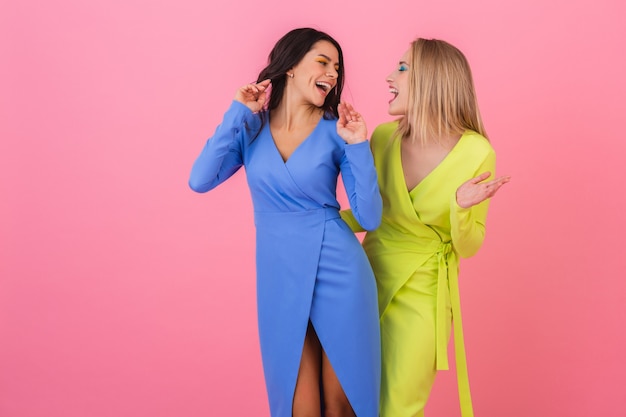 Twee stijlvolle lachende aantrekkelijke vrouwen plezier poseren op roze muur in stijlvolle kleurrijke jurken van blauwe en gele kleur, lente modetrend