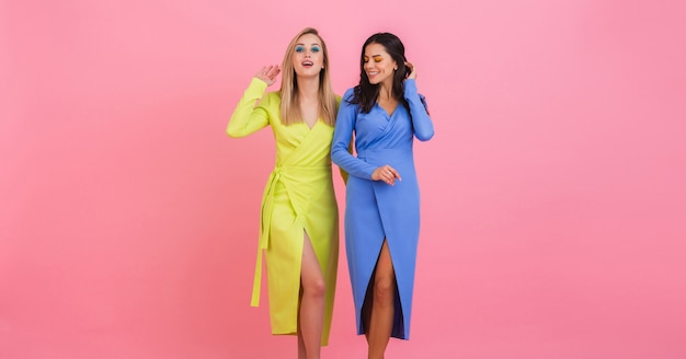 Twee stijlvolle lachende aantrekkelijke vrouwen vrienden poseren op roze muur in stijlvolle kleurrijke jurken van blauwe en gele kleur, lente modetrend
