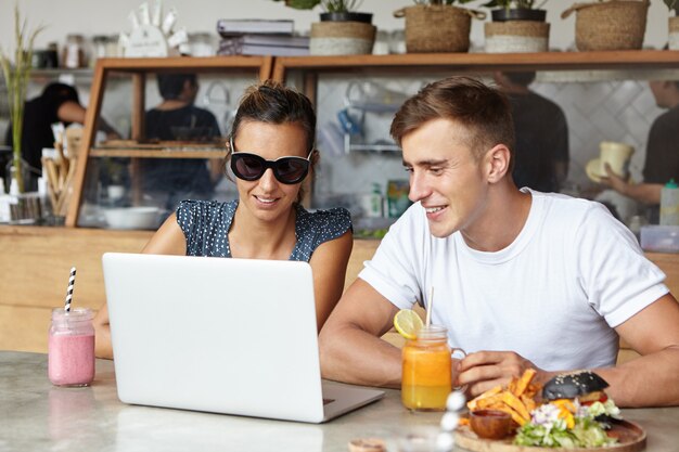 Twee studenten praten, genieten van gratis draadloos internet, laptopcomputer gebruiken, zitten aan tafel met open notebook, verse drankjes en eten tijdens de lunchpauze