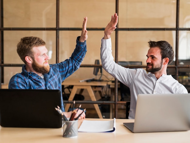 Gratis foto twee succesvolle mannelijke collega die hoge vijf in bureau geeft