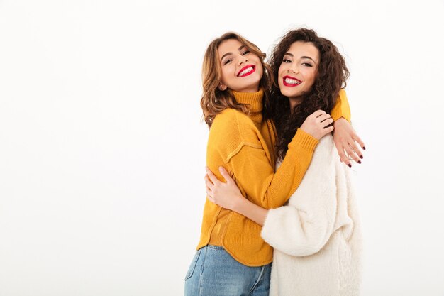 Twee tevreden meisjes in sweaters knuffelen elkaar over witte muur