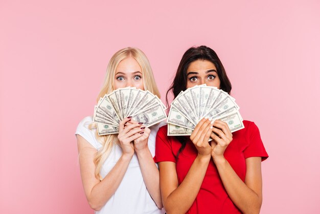Twee verraste vrouwen die zich achter het geld verbergen en de camera over roze bekijken