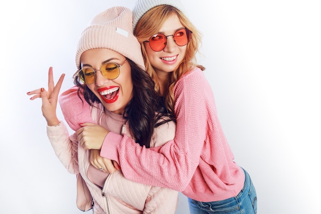 Twee vrolijke jonge vrienden in perfecte stemming met tijd samen op wit isoleren achtergrond. Positieve meisjes met roze knusse truien en pullover, trendy hoed, schattige bril.