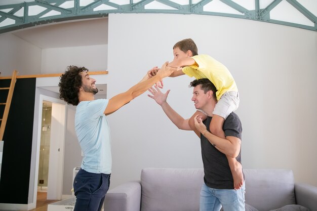 Twee vrolijke vaders en zoon plezier thuis, jongen rijden op mans nek. Zijaanzicht. Familie- en ouderschap concept