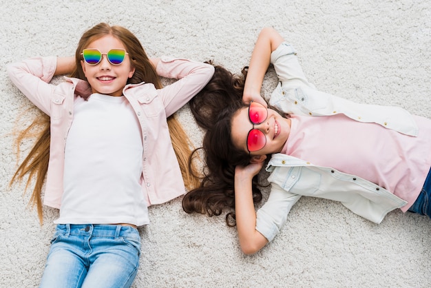 Twee vrouwelijke vrienden die modieuze zonnebril dragen die op wit tapijt liggen