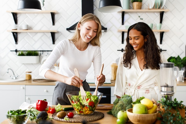 Twee vrouwen van verschillende nationaliteiten glimlachen en koken een salade in de keuken