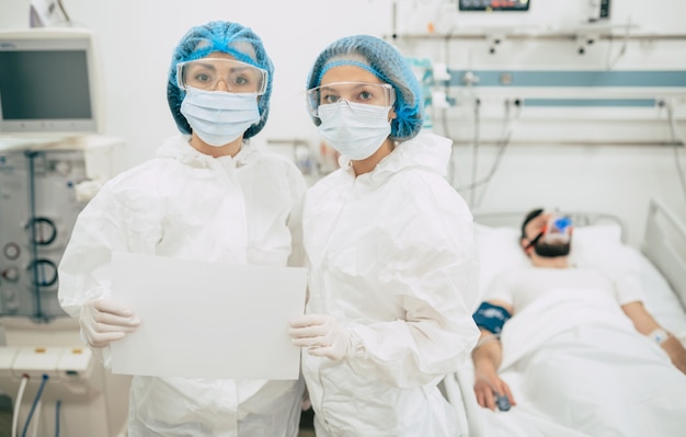 Twee zelfverzekerde artsen in beschermende veiligheidspakken staan op de achtergrond van een patiënt met coronavirus