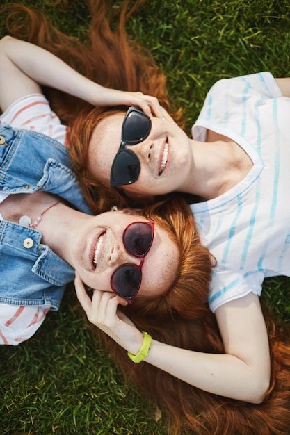 Tweelingzusjes met gember in zonnebrillen die op een zonnige herfstdag op het gras liggen en genieten van de lucht Samen volwassen worden in roodharige waanzin