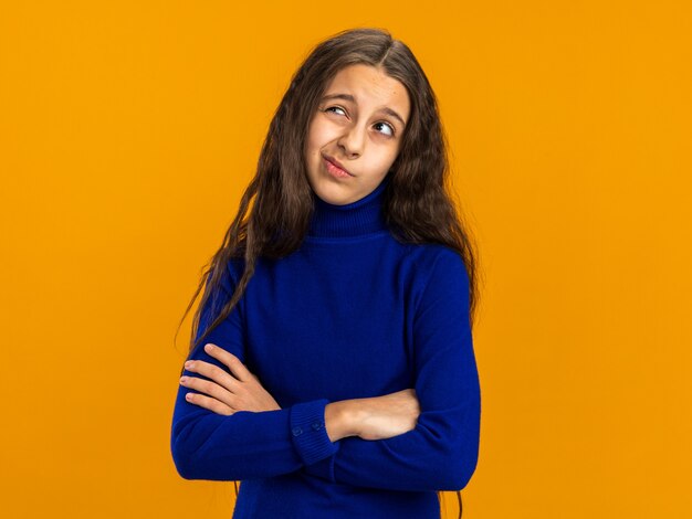 Twijfelachtig tienermeisje permanent met gesloten houding opzoeken geïsoleerd op oranje muur met kopie ruimte