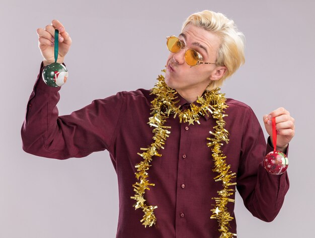 Twijfelachtige jonge blonde man draagt ?? een bril met klatergoud slinger rond de nek met kerstballen kijken naar een geïsoleerd op een witte muur