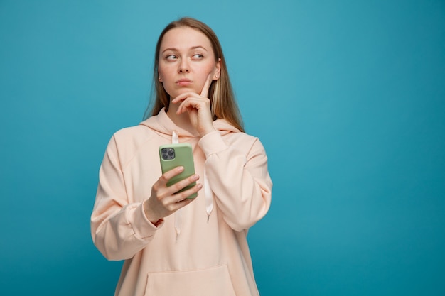 Twijfelachtige jonge blonde vrouw die mobiele telefoon houdt die hand op kin houdt die kant bekijkt