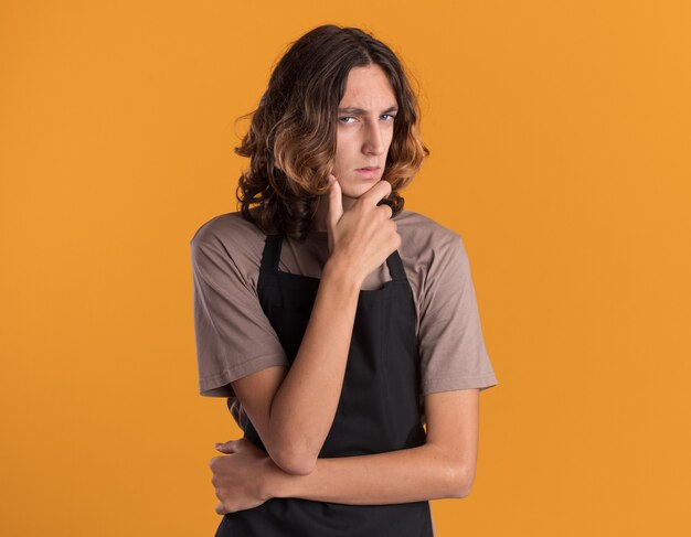 Twijfelachtige jonge knappe kapper in uniform die de hand op de kin houdt en naar de voorkant kijkt geïsoleerd op een oranje muur met kopieerruimte with