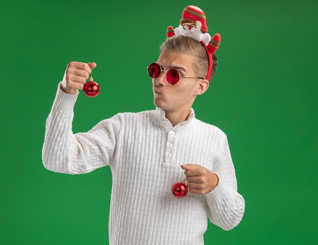 Gratis foto twijfelachtige jonge knappe kerel die de hoofdband van de kerstman met een bril draagt die kerstballen kijkt naar een van hen geïsoleerd op groene muur met kopie ruimte