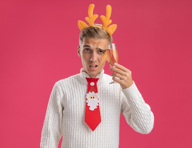 Gratis foto twijfelachtige jonge knappe kerel die rendiergeweien hoofdband en stropdas van de kerstman draagt ?? die glas champagne aanraken hoofd ermee kijken camera geïsoleerd op roze achtergrond