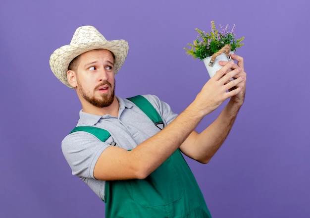 Twijfelachtige jonge knappe Slavische tuinman in uniform en hoed houden en kijken naar bloempot geïsoleerd op paarse muur