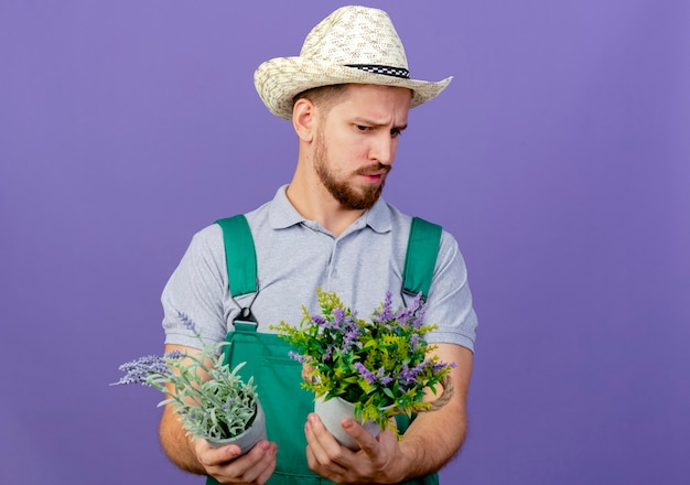 Twijfelachtige jonge knappe Slavische tuinman in uniform en hoed houden en kijken naar bloempotten geïsoleerd op paarse muur met kopie ruimte