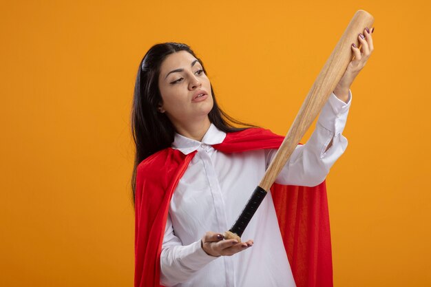 Twijfelachtige jonge superwoman houden en kijken naar honkbalknuppel geïsoleerd op oranje muur