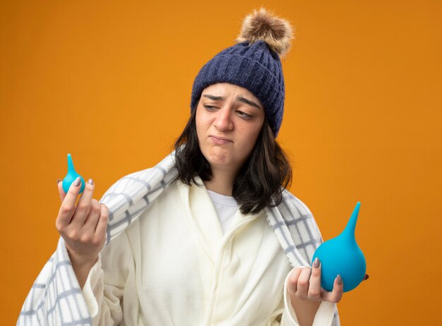 Twijfelachtige jonge zieke vrouw draagt ?? gewaad winter hoed verpakt in plaid bedrijf klysma's kijken naar kleintje geïsoleerd op oranje muur