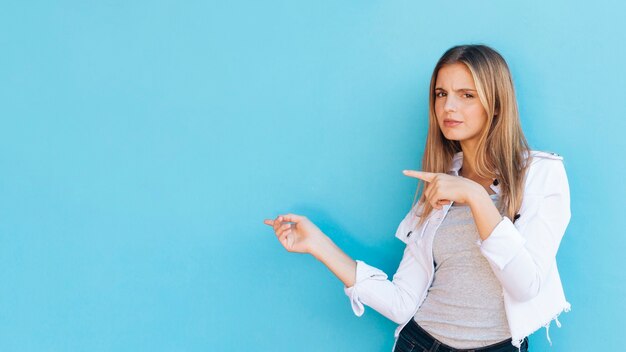 Twijfelachtige mooie blonde jonge vrouw die vingers aan kant richten tegen blauwe achtergrond