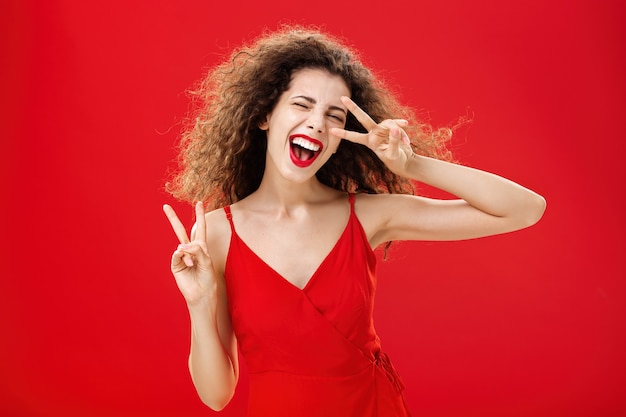 Uitgaande vriendelijke en zorgeloze jonge rebelse vrouw met krullend kapsel in rode stijlvolle jurkshow...