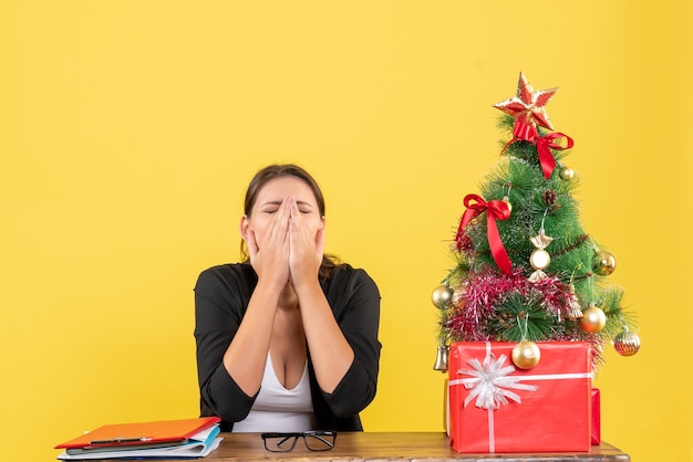 Uitgeput jonge vrouw in pak in de buurt van versierde kerstboom op kantoor op geel