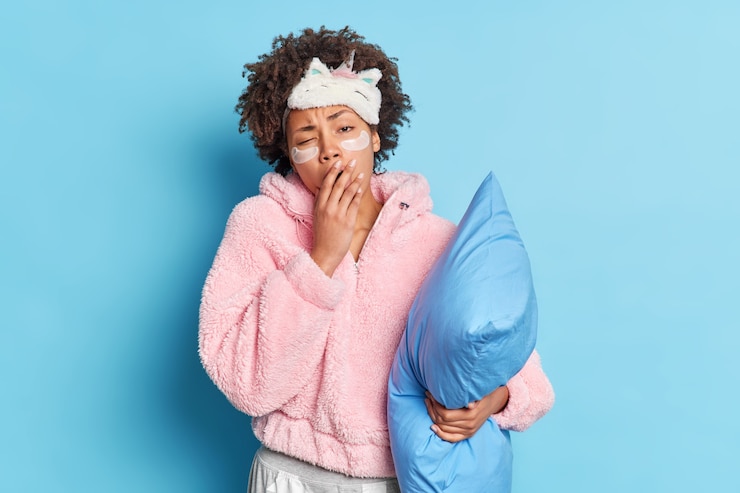 Gratis Foto uitgeput slapeloze vrouw heeft krullend haar gaapt bedekt mond gekleed in nachtkleding houdt kussen vast wil heel vroeg wakker slapen geïsoleerd over blauwe muur