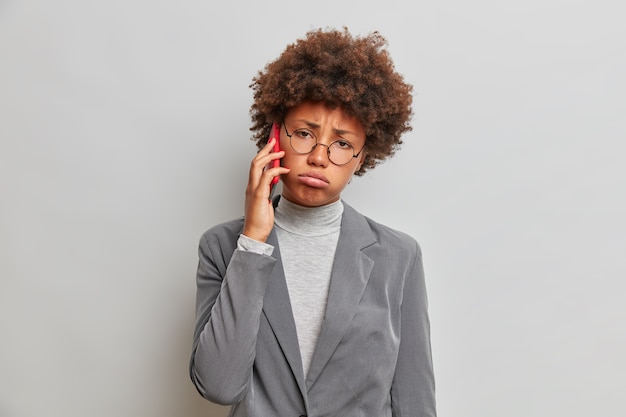 Uitgeput triest zakenvrouw met krullend haar, praat over telefoon, gekleed in trendy formele outfit, heeft een saai gesprek, ziet er ongelukkig uit, zucht van vermoeidheid