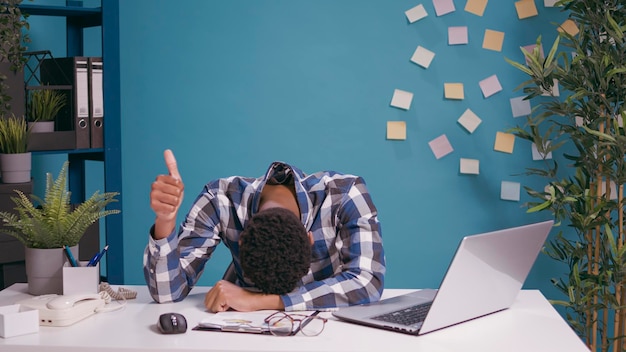 Uitgeputte werknemer die het hoofd op het bureau legt en duimen omhoog laat zien, overwerkt en zich moe voelt. Overwerkte man doet goed gebaar met de hand, wachtend om een pauze te nemen van financieel werk.