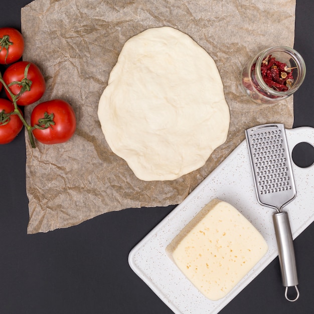 Uitgerold pizzadeeg; tomaten; kaas en gedroogde rode chili met keukengerei op zwarte achtergrond