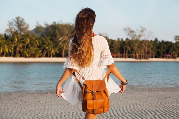 Uitzicht vanaf achterkant mooie vrouw in witte jurk zorgeloos lopen op tropisch strand met lederen rugzak.