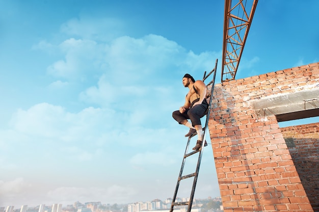 Uitzicht vanaf afstand van bouwer met blote torso en hoed zittend op ladder. leunend op bakstenen muur hoog. Mens die weg kijkt. Blauwe lucht in het zomerseizoen op de achtergrond.