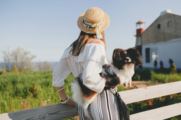 Uitzicht vanaf de achterkant op stijlvolle vrouw op het platteland, met een hond, gelukkige positieve stemming, zomer, strooien hoed, outfit in bohemienstijl,