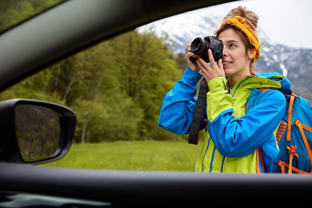 Uitzicht vanaf de auto van professionele jonge vrouw fotograaf neemt foto's op camera, wandelingen op groen veld met berglandschap