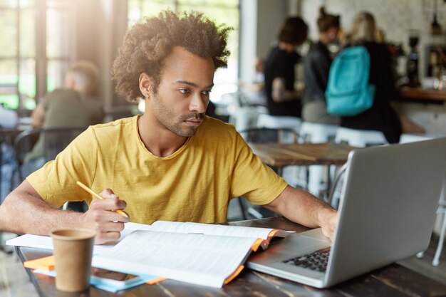 Universiteitsstudent met donkere huid en Afrikaans kapsel zittend in café werken met boeken en notitieblok terwijl ze zich voorbereiden op het examen en de nodige informatie op internet zoeken met een serieuze blik