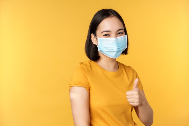 Vaccinatie van covid en gezondheidsconcept Gelukkig Aziatisch meisje dat duimen opsteekt met een medisch maskerpleister op de schouder kreeg een coronavirusvaccin dat op een gele achtergrond is geschoten
