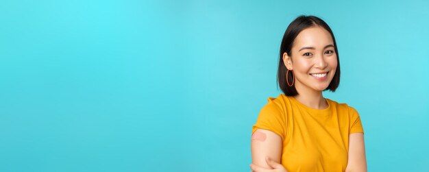 Vaccincampagne van covid19 Jonge mooie gezonde Aziatische vrouw die schouder met pleisterconcept vaccinatie toont die zich over blauwe achtergrond bevindt