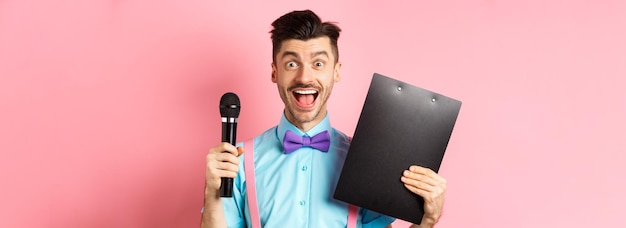 Gratis foto vakantie concept gelukkige jonge man entertainer glimlachend op camera met klembord en microfoon maki