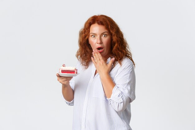 Vakantie, emoties en levensstijlconcept. Geschokte en bezorgde roodharige vrouw van middelbare leeftijd met open mond, hijgend en bezorgd kijkend terwijl ze cake vasthoudt, hoor hoeveel calorieën het heeft.