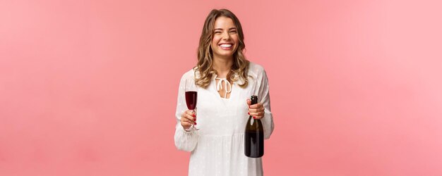 Vakantie lente en feest concept portret van gelukkig en zorgeloos Europese blonde vrouw vieren in witte jurk fles champagne of wijn drinken uit glas en lachen