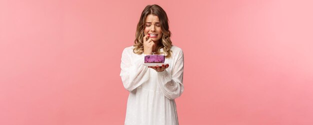 Vakantie lente en feest concept Portret van nerveus blond meisje op dieet lip bijten kan de verleiding niet weerstaan om heerlijke cake te eten kijken naar dessert met aarzelende uitdrukking grimassen bezorgd