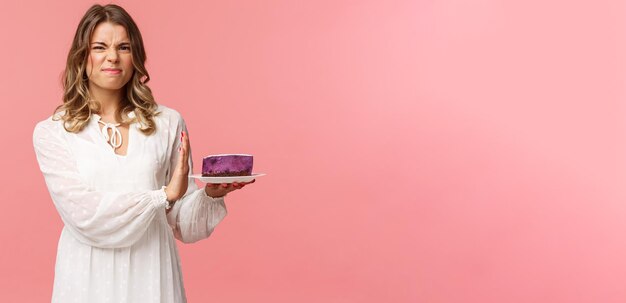 Vakantie lente en feest concept portret van ontevreden en ontevreden blond meisje in witte jurk afwijzen afnemende eten smakeloze taart grimassen en stopbord tonen bij dessert roze achtergrond