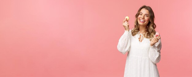 Vakantie lente en feest concept portret van schattig romantisch blond meisje in witte jurk lippen likken als verleidelijk om heerlijk dessert te eten met twee macarons en kijken tevreden roze achtergrond