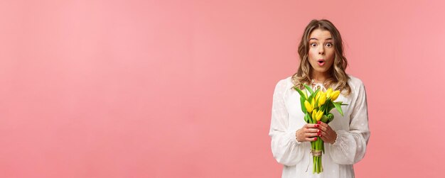 Vakantie schoonheid en lente concept portret van verrast en verbaasd blond meisje in witte jurk met gele tulpen ontvangen bloemen geamuseerd en gelukkig permanent roze achtergrond