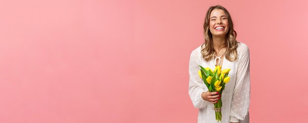 Vakantie schoonheid en lente concept Zorgeloos gelukkig aantrekkelijk blond meisje in witte jurk met gele tulpen en lachen met blije uitdrukking staande roze achtergrond hebben romantische date